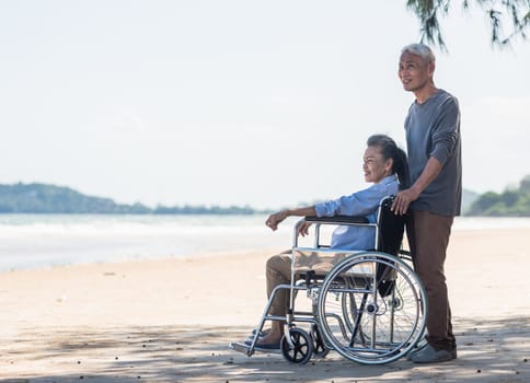 Happy Asian back elderly woman disabled sitting in wheelchair and husband is a wheelchair user on the beach together, summer vacation, Retirement couple concept