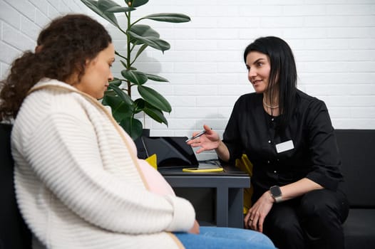 Adult pregnant female patient talking with doctor about test result diagnosis and ultrasound scan of baby at hospital corridor, listening to doctor's prescription. Obstetrics and gynecology concept