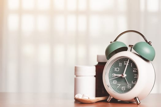 An alarm clock with pills on the wooden spoon and medicine bottles on the table for time management and healthcare concept.