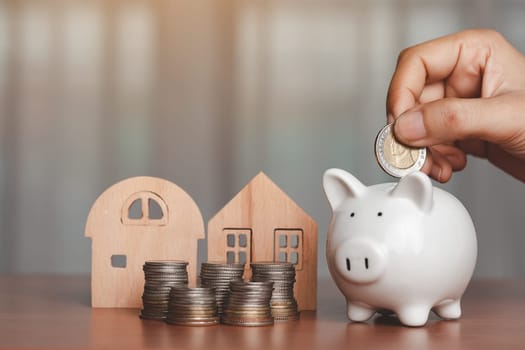 Hand saving a coin into piggy bank with wooden house model and a pile of coins and money on the table for business, finance, saving money and property investment concept.