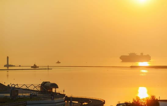 Container ship arrives a port during beautiful sunset along coastline with windmills. The ship sails to the lighthouse