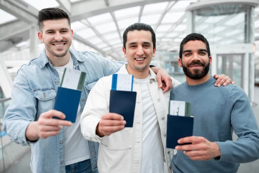 Globetrotters Unite. Three Happy Guys Travelers Embracing At Airport Indoor, Advertising Cheap Tickets Offer Showing Their Passports And Boarding Passes To Camera. Easy Travel And Transportation