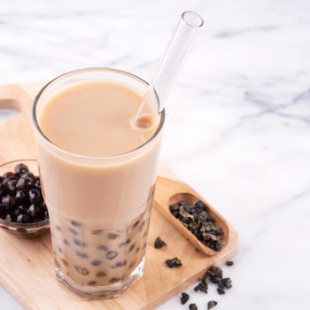 Tapioca pearl ball bubble milk tea, popular Taiwan drink, in drinking glass with straw on marble white table and wooden tray, close up, copy space.