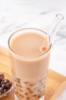 Tapioca pearl ball bubble milk tea, popular Taiwan drink, in drinking glass with straw on marble white table and wooden tray, close up, copy space.