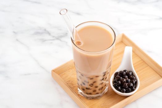 Tapioca pearl ball bubble milk tea, popular Taiwan drink, in drinking glass with straw on marble white table and wooden tray, close up, copy space.