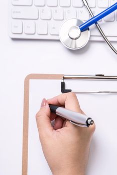 Female doctor writing a medical record case over clipboard on white working table with stethoscope, computer keyboard. Top view, flat lay, copy space