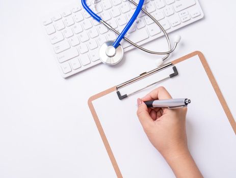 Female doctor writing a medical record case over clipboard on white working table with stethoscope, computer keyboard. Top view, flat lay, copy space