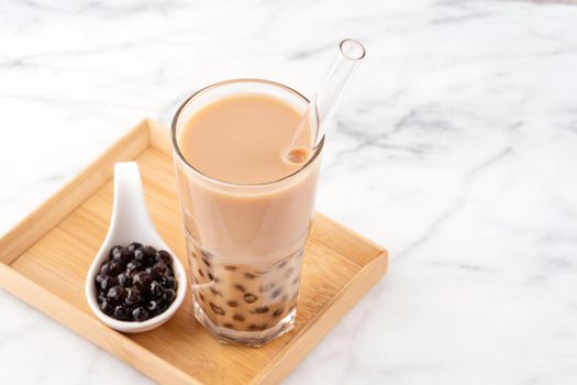 Tapioca pearl ball bubble milk tea, popular Taiwan drink, in drinking glass with straw on marble white table and wooden tray, close up, copy space.