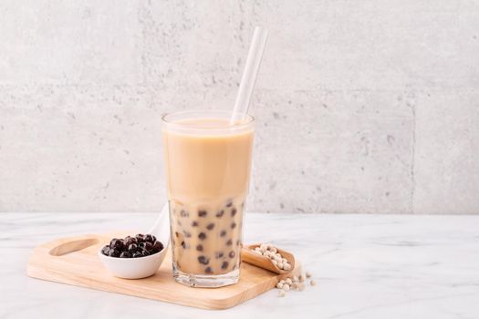 Tapioca pearl ball bubble milk tea, popular Taiwan drink, in drinking glass with straw on marble white table and wooden tray, close up, copy space.