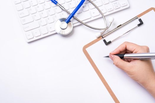 Female doctor writing a medical record case over clipboard on white working table with stethoscope, computer keyboard. Top view, flat lay, copy space