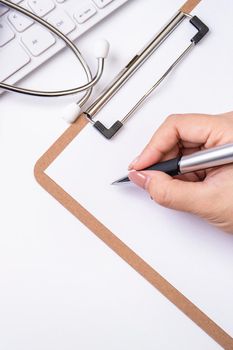 Female doctor writing a medical record case over clipboard on white working table with stethoscope, computer keyboard. Top view, flat lay, copy space