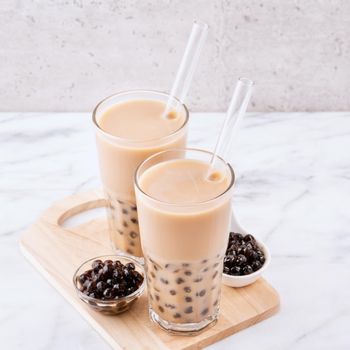 Tapioca pearl ball bubble milk tea, popular Taiwan drink, in drinking glass with straw on marble white table and wooden tray, close up, copy space.