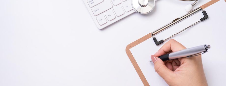 Female doctor writing a medical record case over clipboard on white working table with stethoscope, computer keyboard. Top view, flat lay, copy space