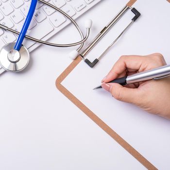 Female doctor writing a medical record case over clipboard on white working table with stethoscope, computer keyboard. Top view, flat lay, copy space