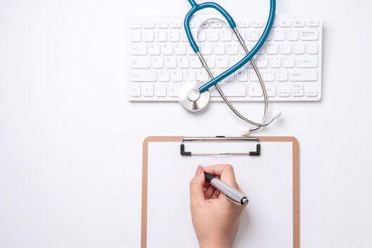 Female doctor writing a medical record case over clipboard on white working table with stethoscope, computer keyboard. Top view, flat lay, copy space