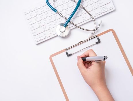 Female doctor writing a medical record case over clipboard on white working table with stethoscope, computer keyboard. Top view, flat lay, copy space
