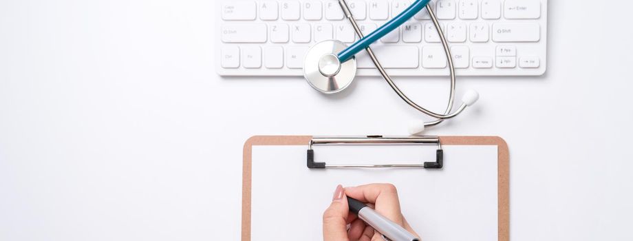 Female doctor writing a medical record case over clipboard on white working table with stethoscope, computer keyboard. Top view, flat lay, copy space