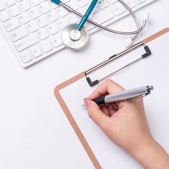 Female doctor writing a medical record case over clipboard on white working table with stethoscope, computer keyboard. Top view, flat lay, copy space