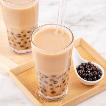Tapioca pearl ball bubble milk tea, popular Taiwan drink, in drinking glass with straw on marble white table and wooden tray, close up, copy space.