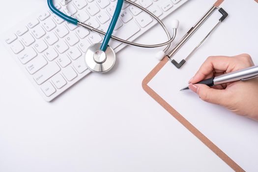 Female doctor writing a medical record case over clipboard on white working table with stethoscope, computer keyboard. Top view, flat lay, copy space
