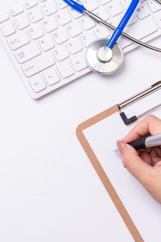 Female doctor writing a medical record case over clipboard on white working table with stethoscope, computer keyboard. Top view, flat lay, copy space
