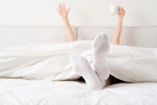 Female hand holding cup of coffee from under a blanket in bed. Woman waking up in the morning.