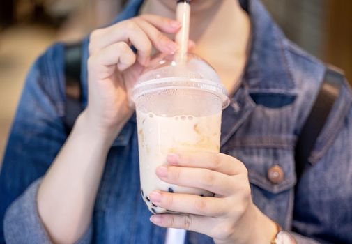 Young girl in denim jacket is drinking brown sugar flavored tapioca pearl bubble milk tea with glass straw in night market of Taiwan, close up, bokeh