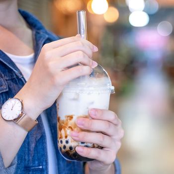 Young girl in denim jacket is drinking brown sugar flavored tapioca pearl bubble milk tea with glass straw in night market of Taiwan, close up, bokeh