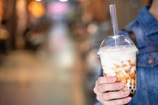 Young girl in denim jacket is drinking brown sugar flavored tapioca pearl bubble milk tea with glass straw in night market of Taiwan, close up, bokeh