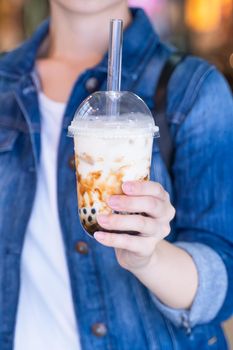 Young girl in denim jacket is drinking brown sugar flavored tapioca pearl bubble milk tea with glass straw in night market of Taiwan, close up, bokeh