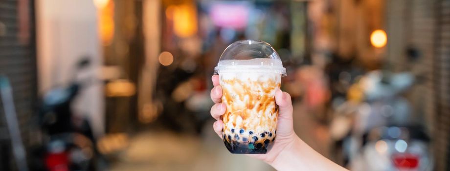 Young girl is holding and showing a cup of brown sugar flavored tapioca pearl bubble milk tea in night market of Taiwan background, close up, bokeh