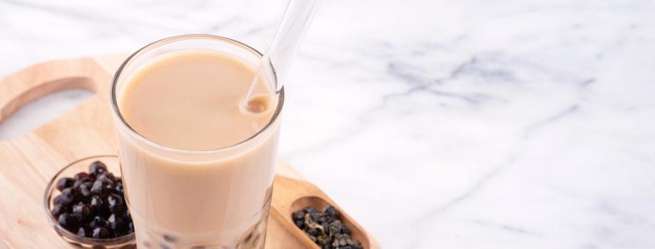 Tapioca pearl ball bubble milk tea, popular Taiwan drink, in drinking glass with straw on marble white table and wooden tray, close up, copy space.