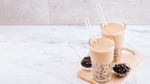 Tapioca pearl ball bubble milk tea, popular Taiwan drink, in drinking glass with straw on marble white table and wooden tray, close up, copy space.