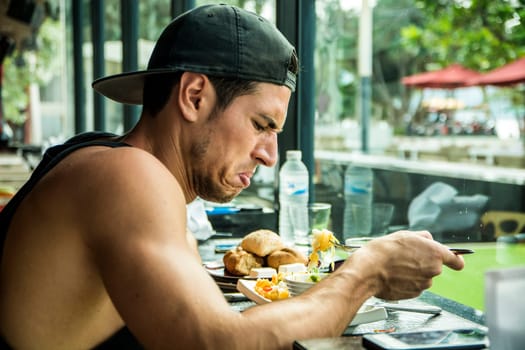 Side view of young man in cap with disgust face expression sitting at table near window and eating food in cafe