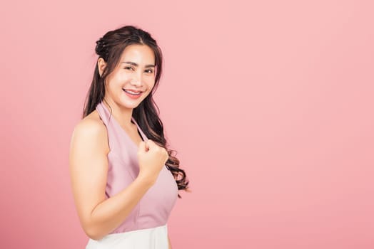 Happy Asian portrait beautiful cute young woman stand makes raised fists up celebrating her winning success gesture, studio shot isolated pink background, Thai female excited say yes with copy space