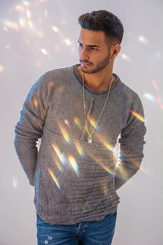 Handsome tattooed young man wearing grey sweater, standing against light background in studio shot with specks of light floating