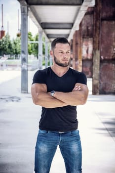 Handsome muscular athletic man in city park during the day, wearing black t-shirt, with arms crossed on chest