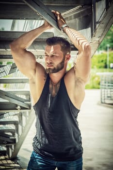 Handsome muscular athletic man in city park under metal stairs, during the day, wearing black tank top