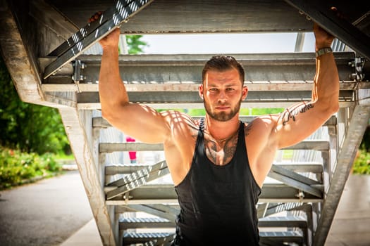 Handsome muscular athletic man in city park under metal stairs, during the day, wearing black tank top