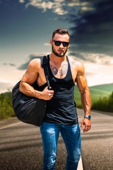 Handsome muscular athletic man walking on empty road at sunset, wearing black t-shirt, carrying a big bag
