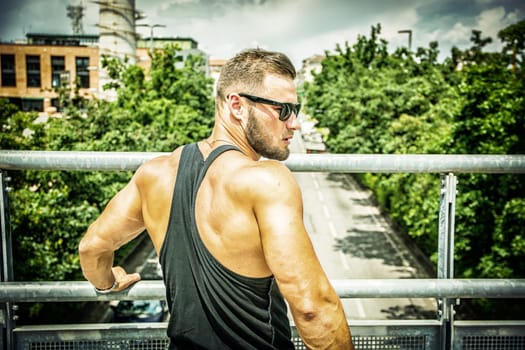 Handsome muscular athletic man in city park during the day, above a street, wearing black tank top and sunglasses