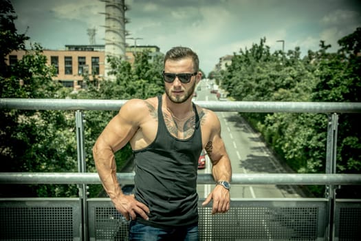 Handsome muscular athletic man in city park during the day, above a street, wearing black tank top and sunglasses