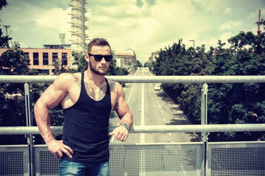 Handsome muscular athletic man in city park during the day, above a street, wearing black tank top and sunglasses