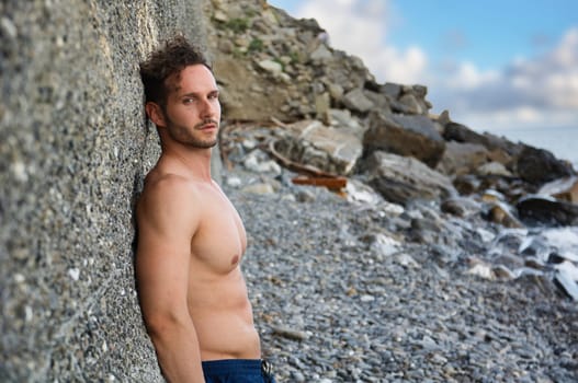 Attractive muscular shirtless athletic man standing on rock next to water by sea or ocean shore, looking at camera in a cloudy summer day