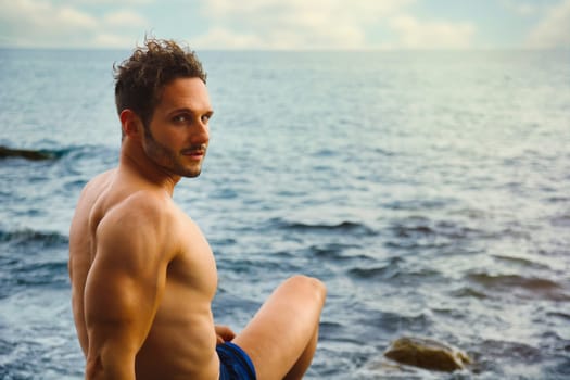 Attractive muscular shirtless athletic man sitting next to water by sea or ocean shore, looking at camera in a cloudy summer day