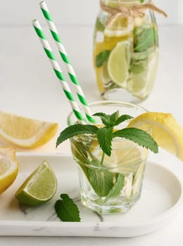 Lemonade in a transparent glass with lemon, lime, rosemary sprigs and mint leaves on a white background