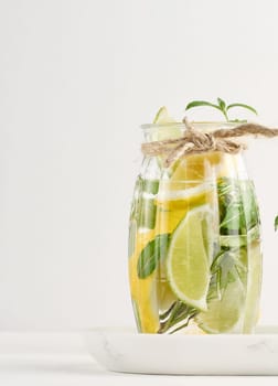 Lemonade in a transparent glass with lemon, lime, rosemary sprigs and mint leaves on a white background