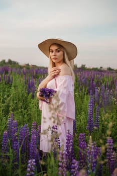 A beautiful woman in a straw hat walks in a field with purple flowers. A walk in nature in the lupin field.