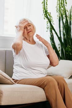 Elderly woman severe pain in the head sitting on the couch, health problems in old age, poor quality of life. Grandmother with gray hair holding her head, migraine and high blood pressure. High quality photo