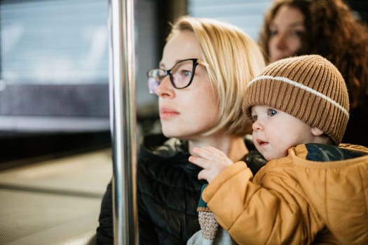 Mother carries her child while standing and holding on to the bus. Mom holding her infant baby boy in her arms while riding in a public transportation. Cute toddler boy traveling with his mother